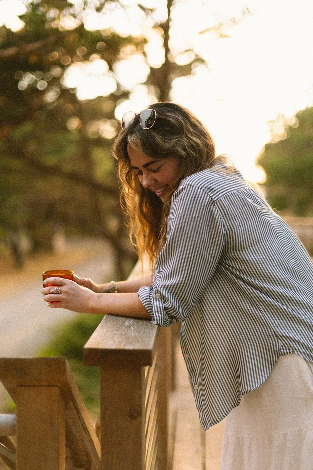 A woman with long, wavy hair wearing sunglasses on her head and a striped shirt, smiling while looking down at a mug she is holding, leaning on a wooden railing outdoors during sunset.
