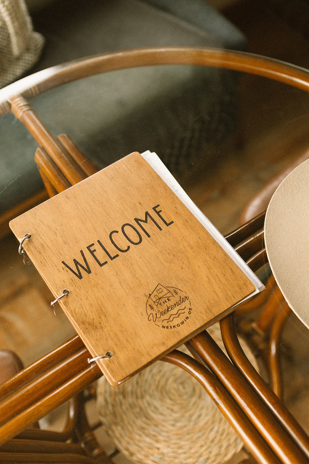 A wooden welcome sign with the words 'WELCOME' and 'The Weekend' on it, placed on top of a glass table next to woven chairs.