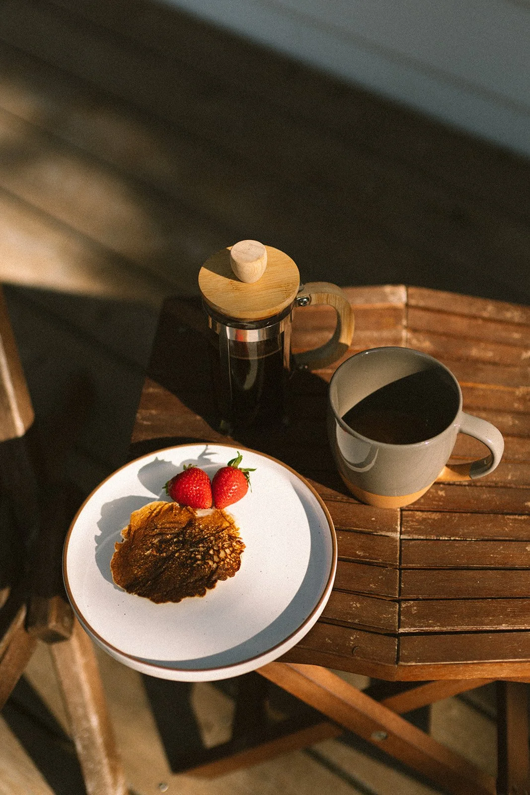 A wooden table with a white plate holding two strawberries and a pancake, next to a mug of black coffee and a French press on a dark background.