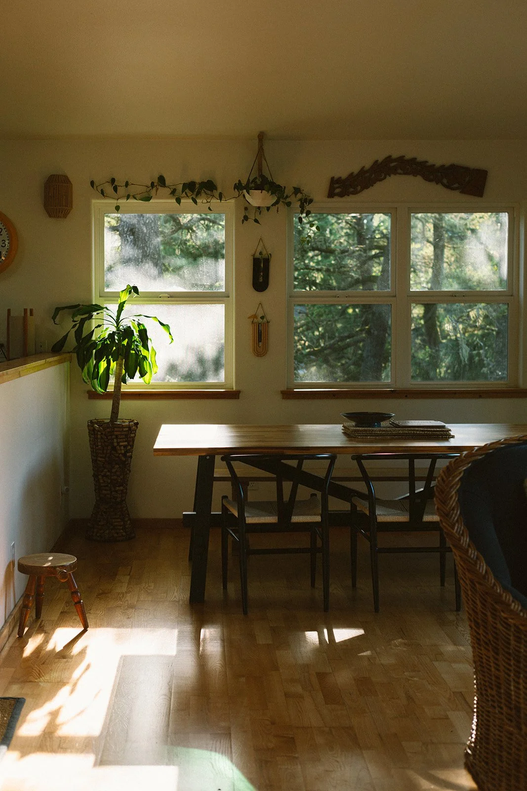 Sunlit dining area with wooden table, black chairs, large windows showing green trees outside, potted plant, hanging decorative items, and natural wood flooring.