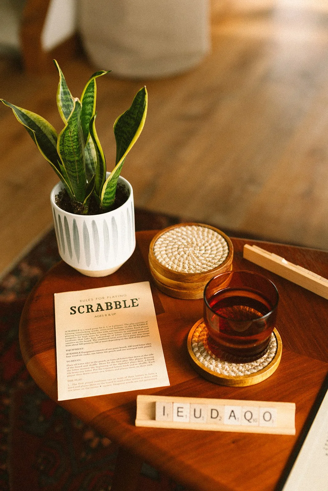 A wooden table with a potted snake plant, a Scrabble game sheet, a glass of dark liquid on a coaster, a woven coaster, and wooden Scrabble tiles spelling 'IEUDAQO' at the bottom.