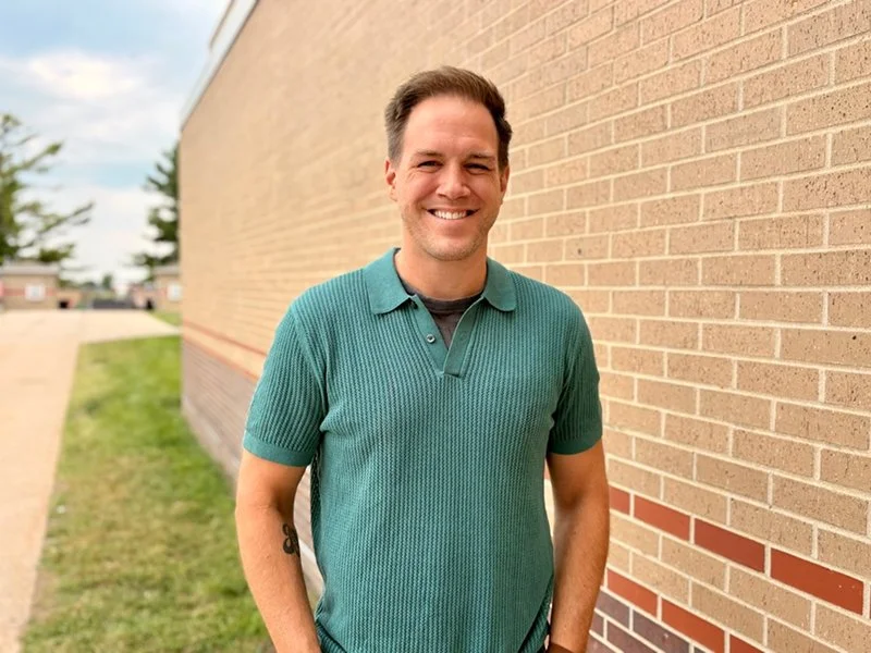 Smiling man in teal polo shirt standing outdoors next to a brick wall