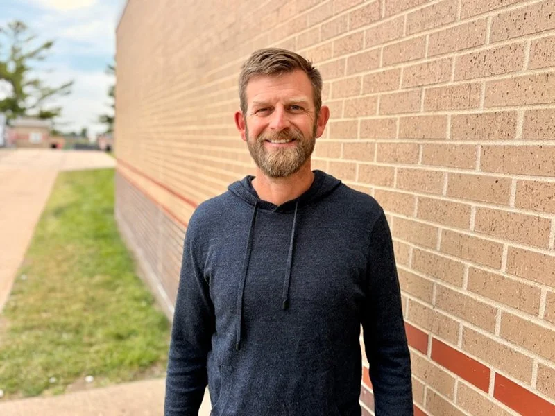 Smiling middle-aged man with a beard, wearing a dark hoodie, standing against a brick wall outdoors.