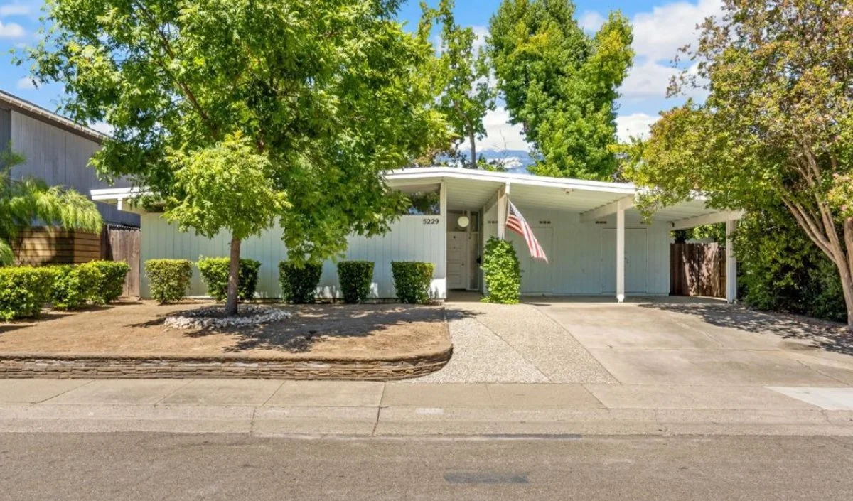 WHte mid century modern home with carport. Big green trees. American flag. Aggregate driveway.