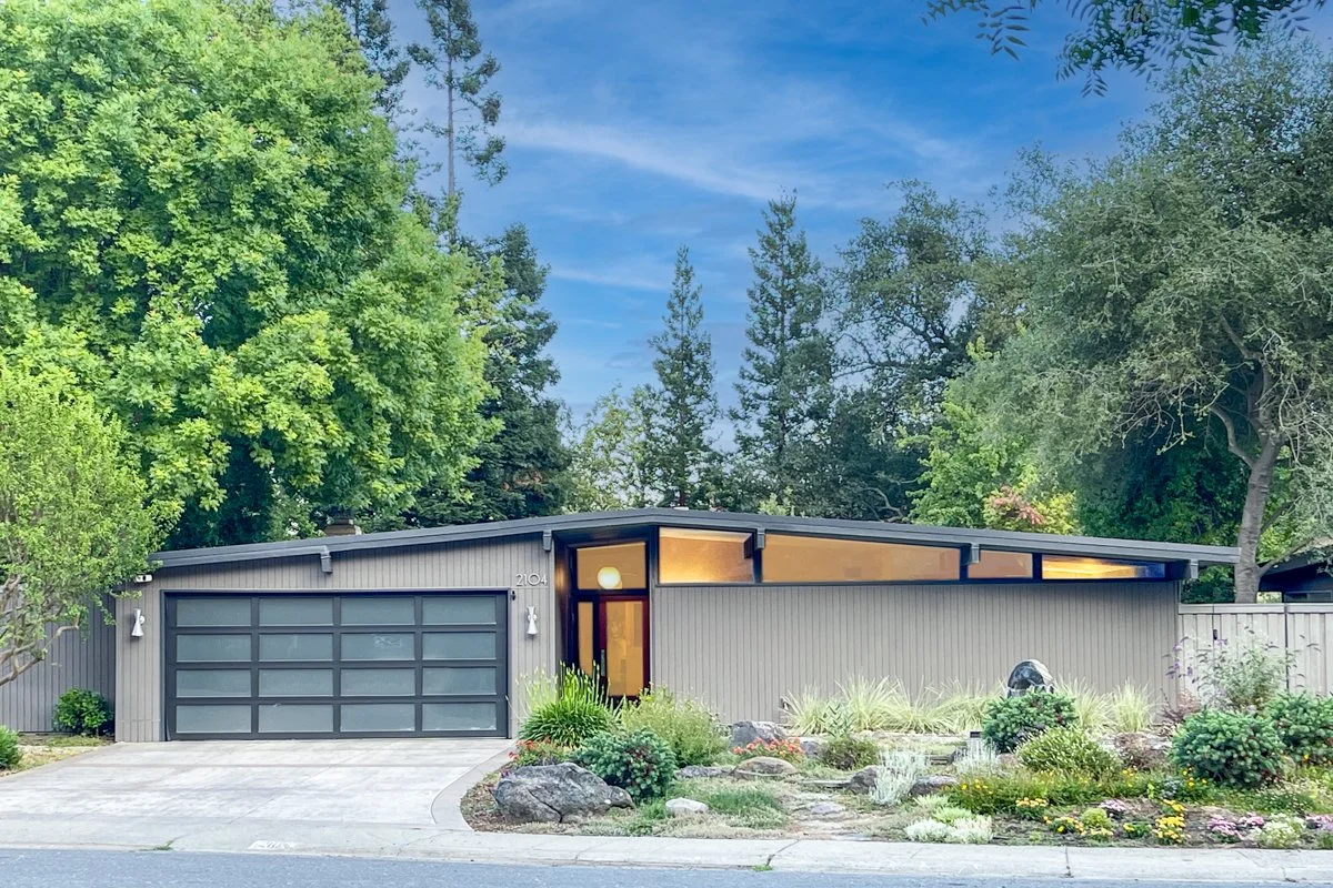 Mid century modern home. Grey paint and dark gray trim. Translucent 2 car garage door. Lights on with clerestory windows.