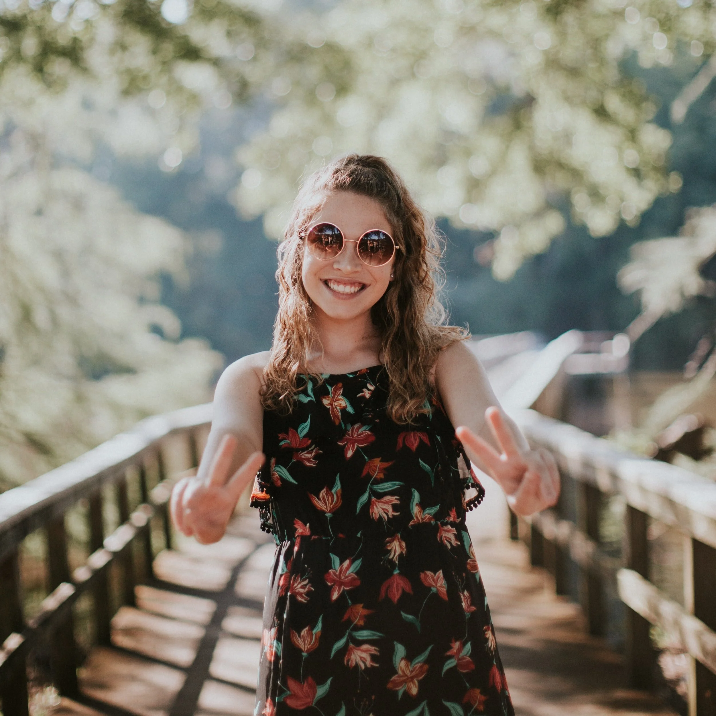 Woman with curly hair wearing sunglasses and a floral dress standing on a bridge, smiling and making peace signs with both hands.