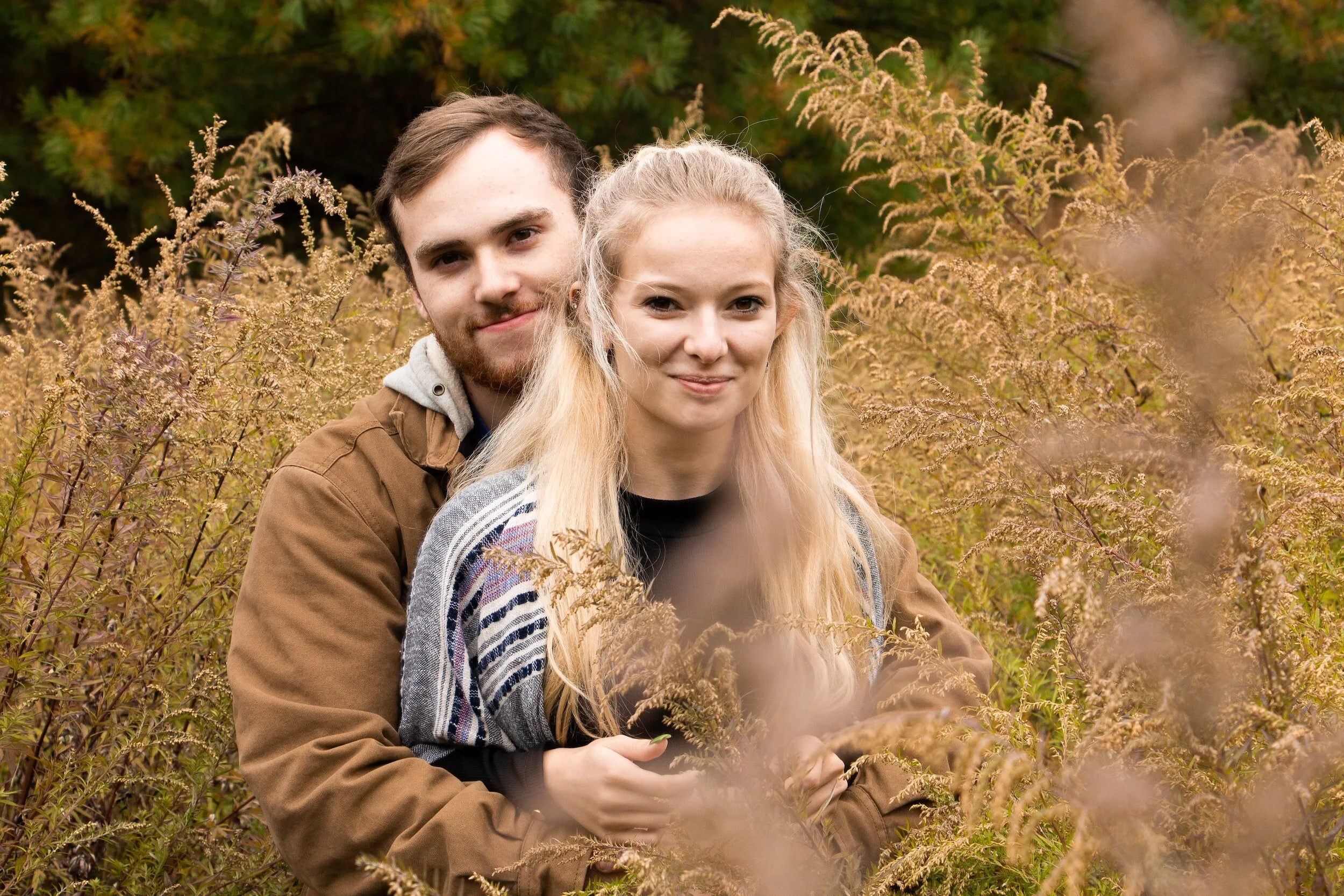 A couple embracing in a field with tall grass and plants, with green trees in the background.