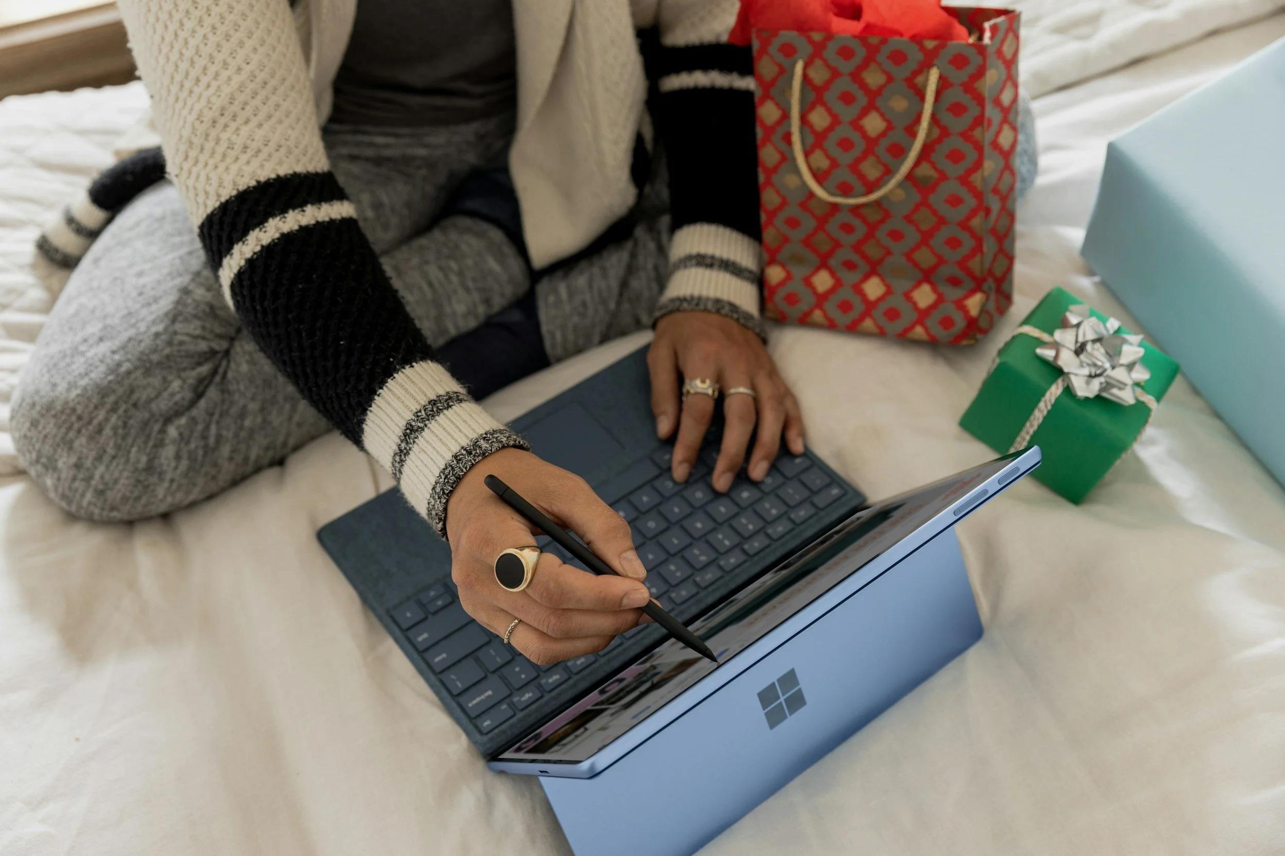 Person using a stylus on a Microsoft Surface laptop with gift bags and wrapped presents nearby on a bed.