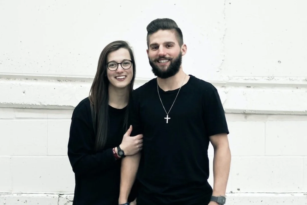A smiling man and woman standing together against a white wall. The woman is wearing glasses and a black top, and the man is wearing a black shirt with a cross necklace.