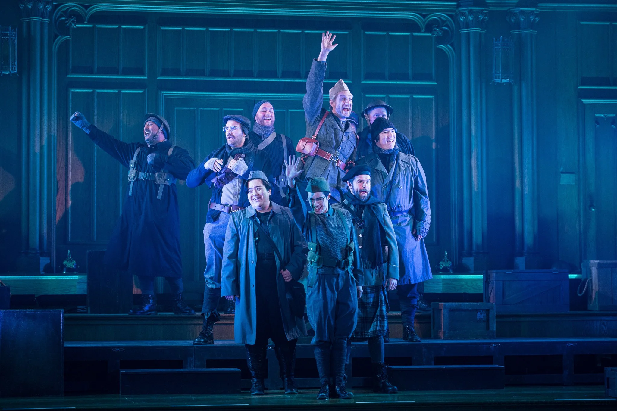 The cast of All Is Calm on stage, dressed in military and winter clothing, celebrating with some raising their hands and smiling. Wood paneling in the background.