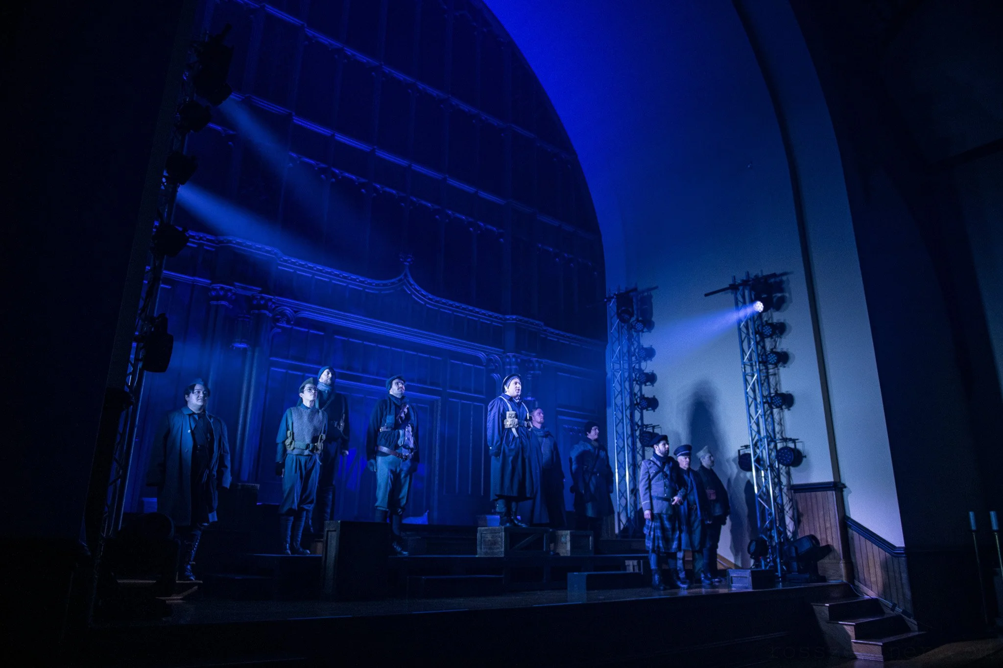 A theatrical stage with thirteen people standing in WWI military dress on risers, illuminated by blue lighting, performing All Is Calm.