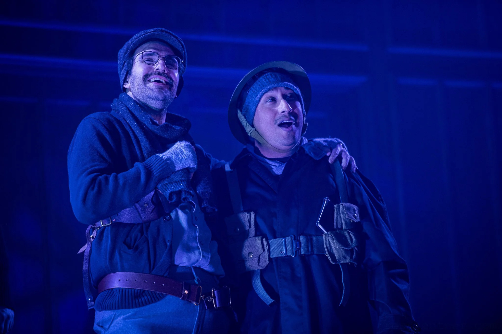 Two men in All is Calm are smiling in celebration, one has his arm around the other. Both are dressed warmly with hats, gloves, and WWI uniforms.