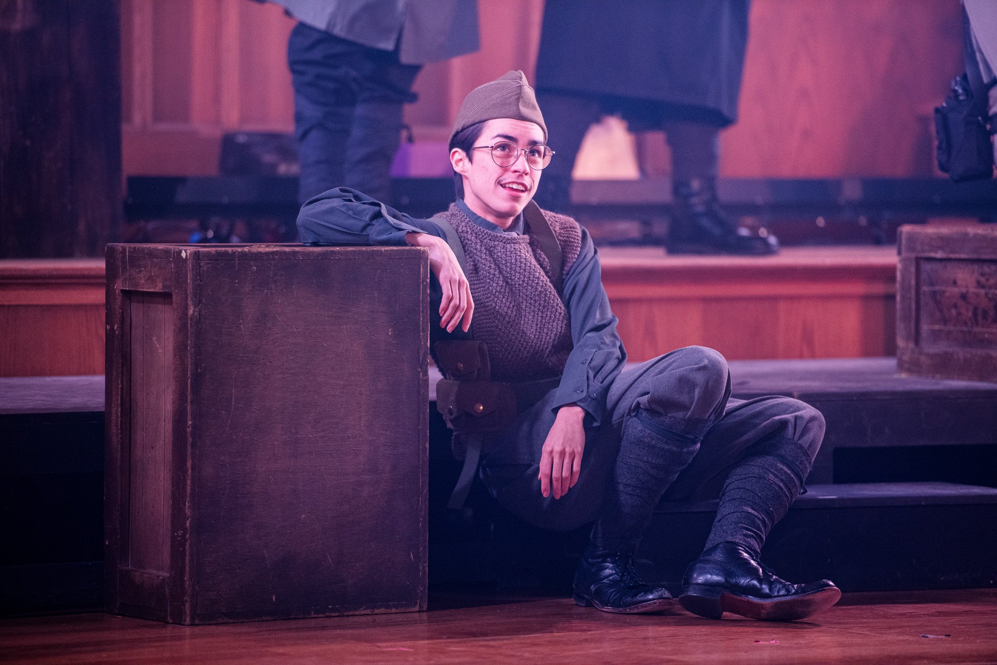 A young man dressed in WWI military attire, sitting on the stage floor with one arm resting on a wooden crate. He is wearing glasses, a knit vest over a long-sleeve shirt, dark trousers, puttees, and black boots, with a garrison cap on his head. 