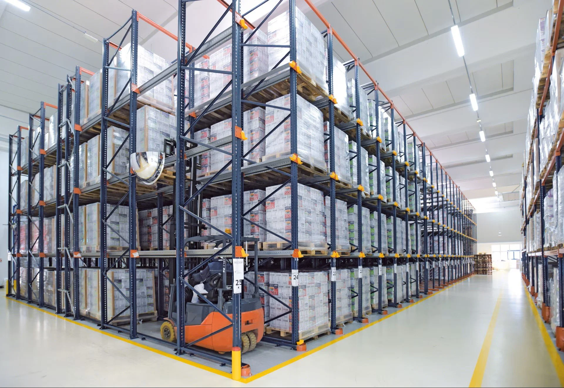 Warehouse with tall blue metal shelving units filled with boxed goods, and a small orange forklift parked near the shelves.