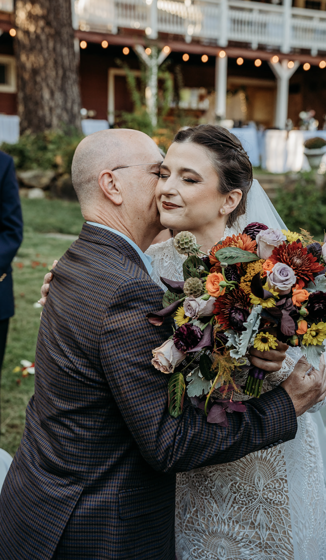 An older man embracing a bride, who holds a colorful bouquet, during a wedding celebration outdoors.