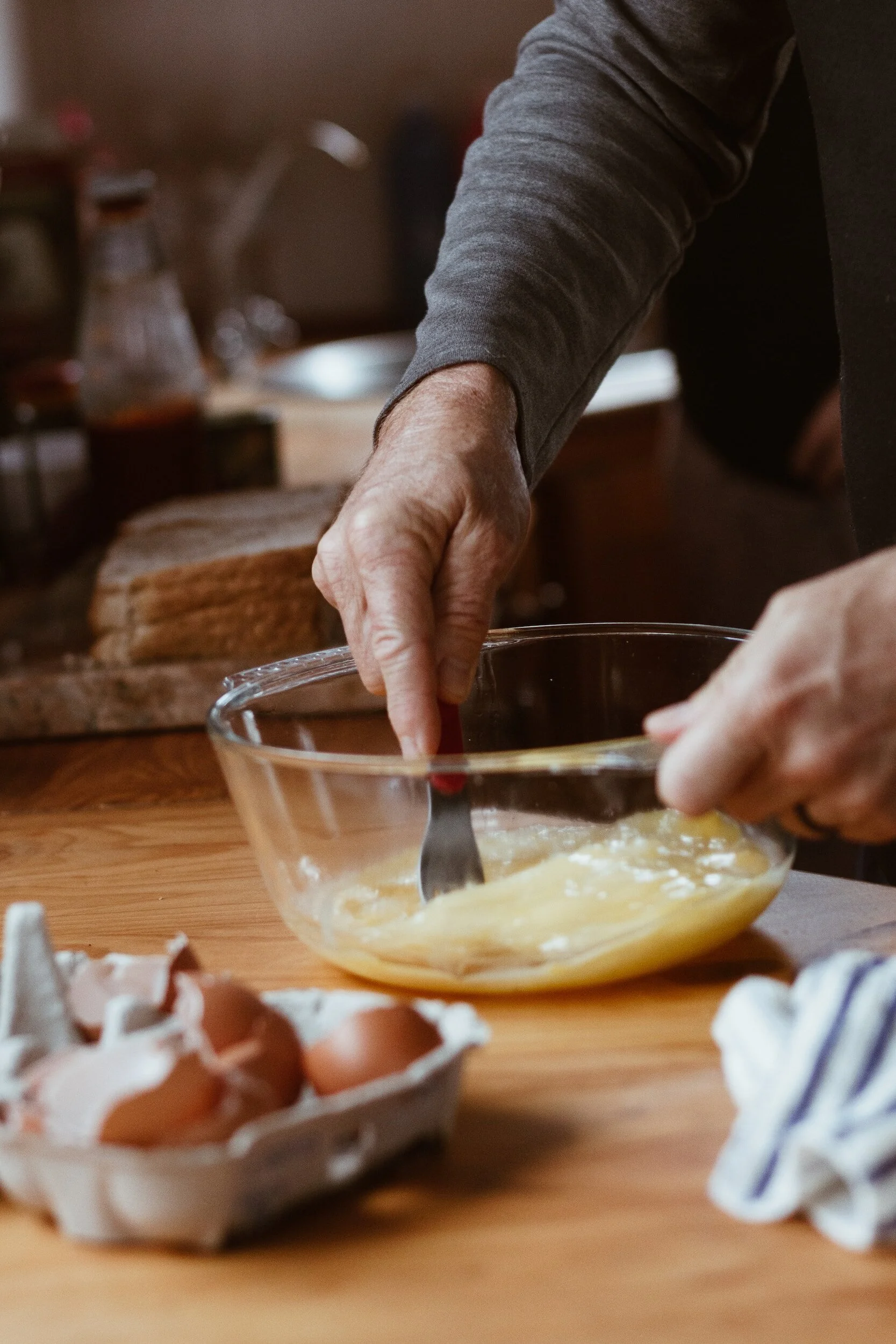 A person whisking batter in a glass bowl on a wooden kitchen counter, with a carton of eggs nearby.