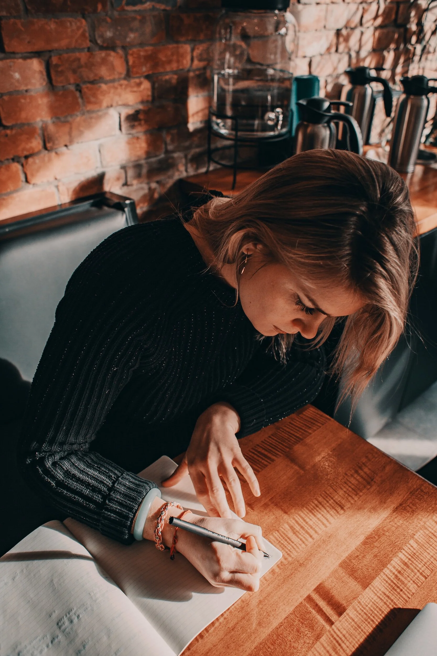 A woman with blonde hair sitting at a wooden table, writing in a notebook with a pen, in a room with a brick wall background and coffee makers on the counter.