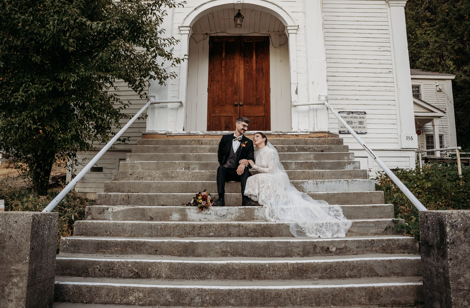 A bride and groom sitting on the steps of a white wooden church, looking at each other lovingly, with a bouquet of flowers placed on the step.