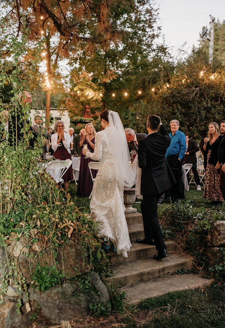 Bride and groom at outdoor wedding ceremony with guests, trees, string lights, and a fountain.