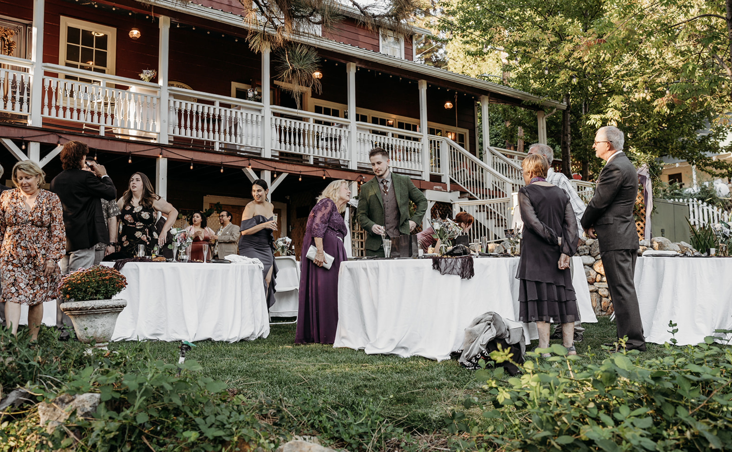 Group of people socializing at an outdoor event with tables covered in white tablecloths, set against a two-story house with a porch, surrounded by greenery and trees.