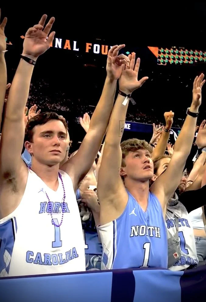 College basketball fans wearing North Carolina jerseys raising their arms during a game in a packed arena.