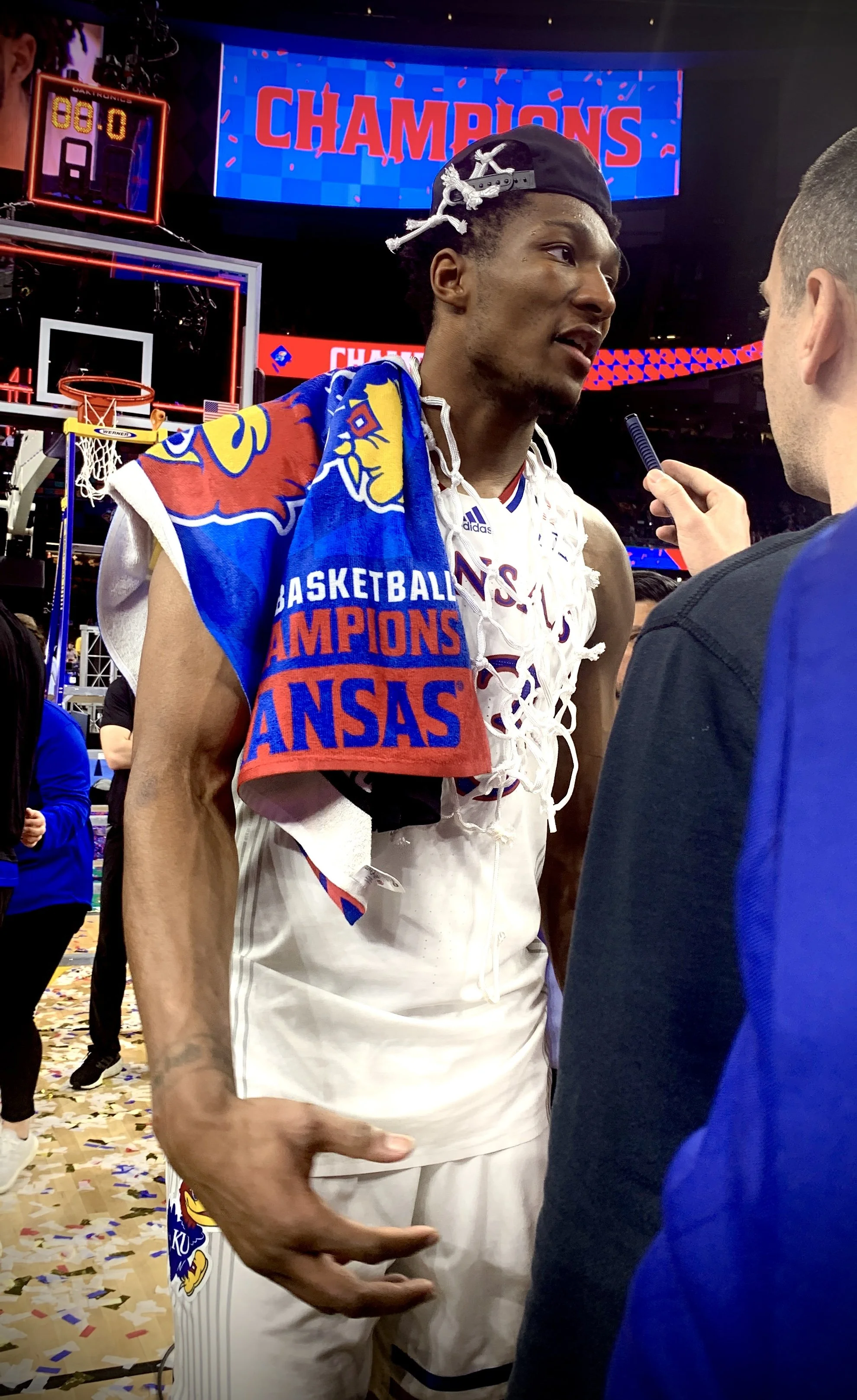 A basketball player being interviewed after winning the championship, with a towel over his shoulder and the word 'CHAMPIONS' on a large screen behind him.