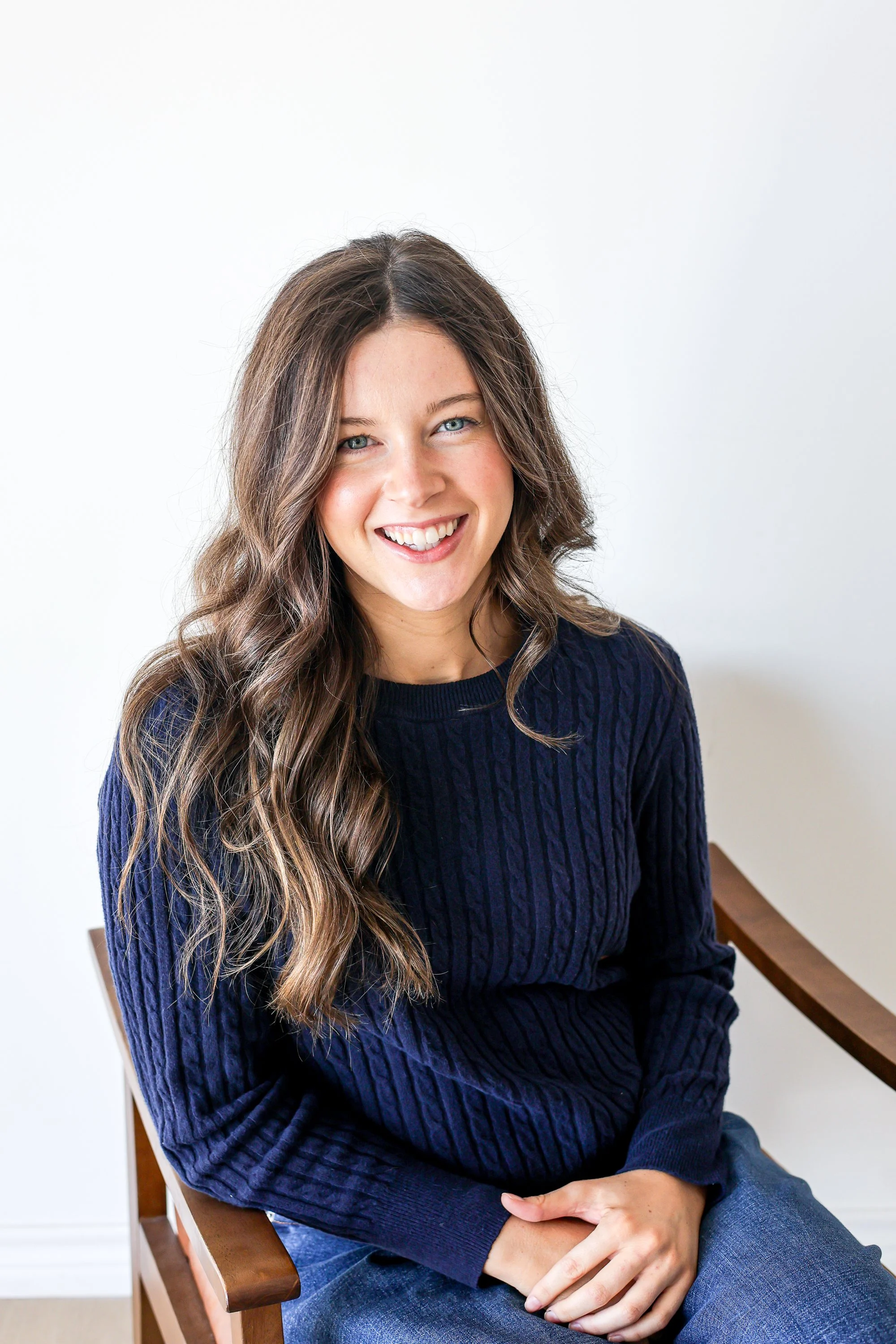 A young woman with long, wavy brown hair and blue eyes smiling while sitting on a wooden chair against a plain white wall, wearing a navy blue cable knit sweater.