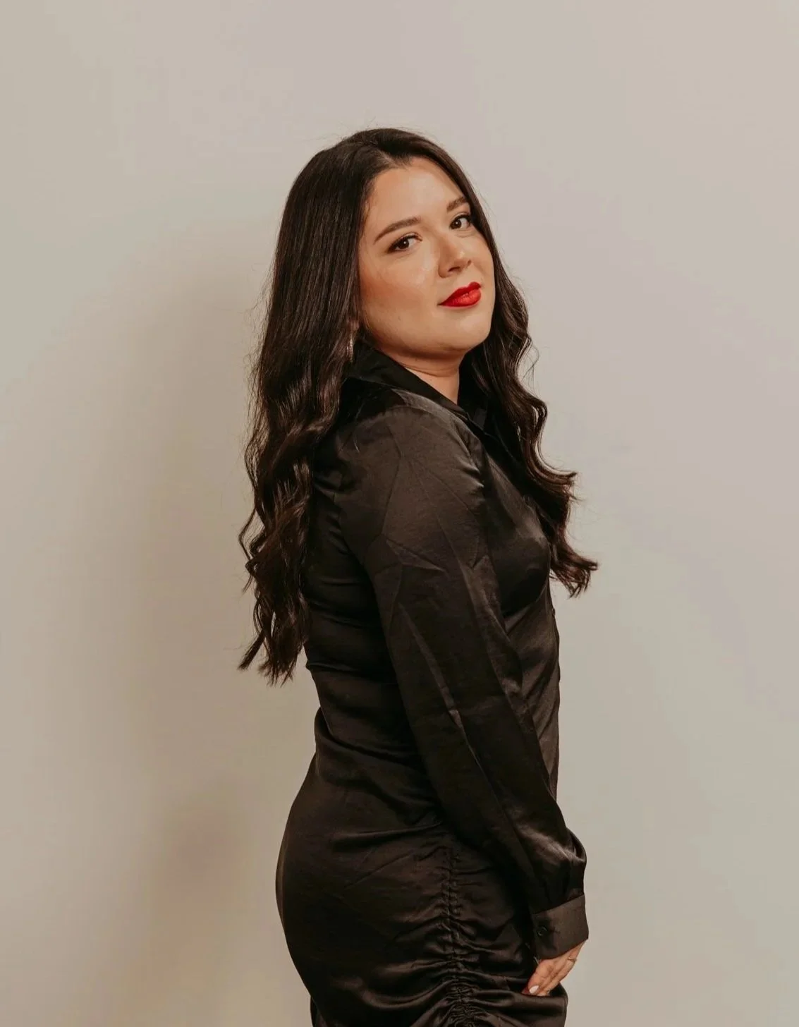A woman with long dark wavy hair wearing a black satin dress standing against a plain light-colored wall. AP Luxe Stylist in Mill Valley luxury hair stylist in Marin County