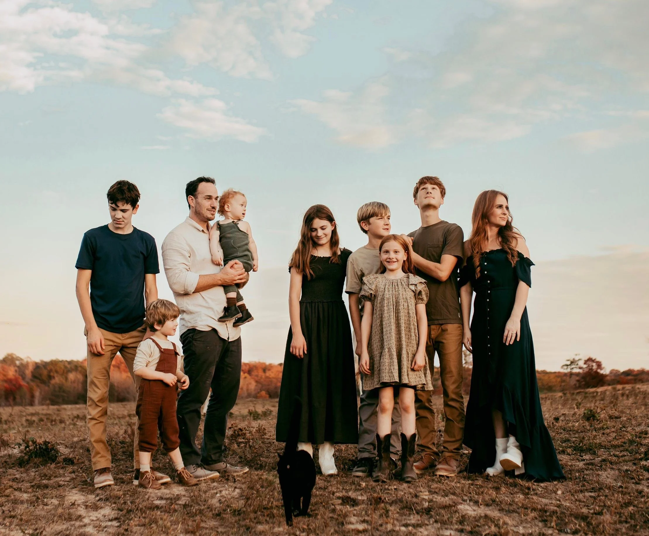 A large family standing outdoors on a field during sunset, including adults and children, dressed in casual and semi-formal clothing, with a dog sitting in front.