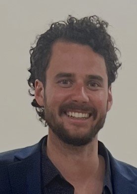 Fresh vegetables including cherry tomatoes, zucchinis, potatoes, sweet potatoes, corn, leafy greens, basil, broccoli, and onions arranged on a white surface.