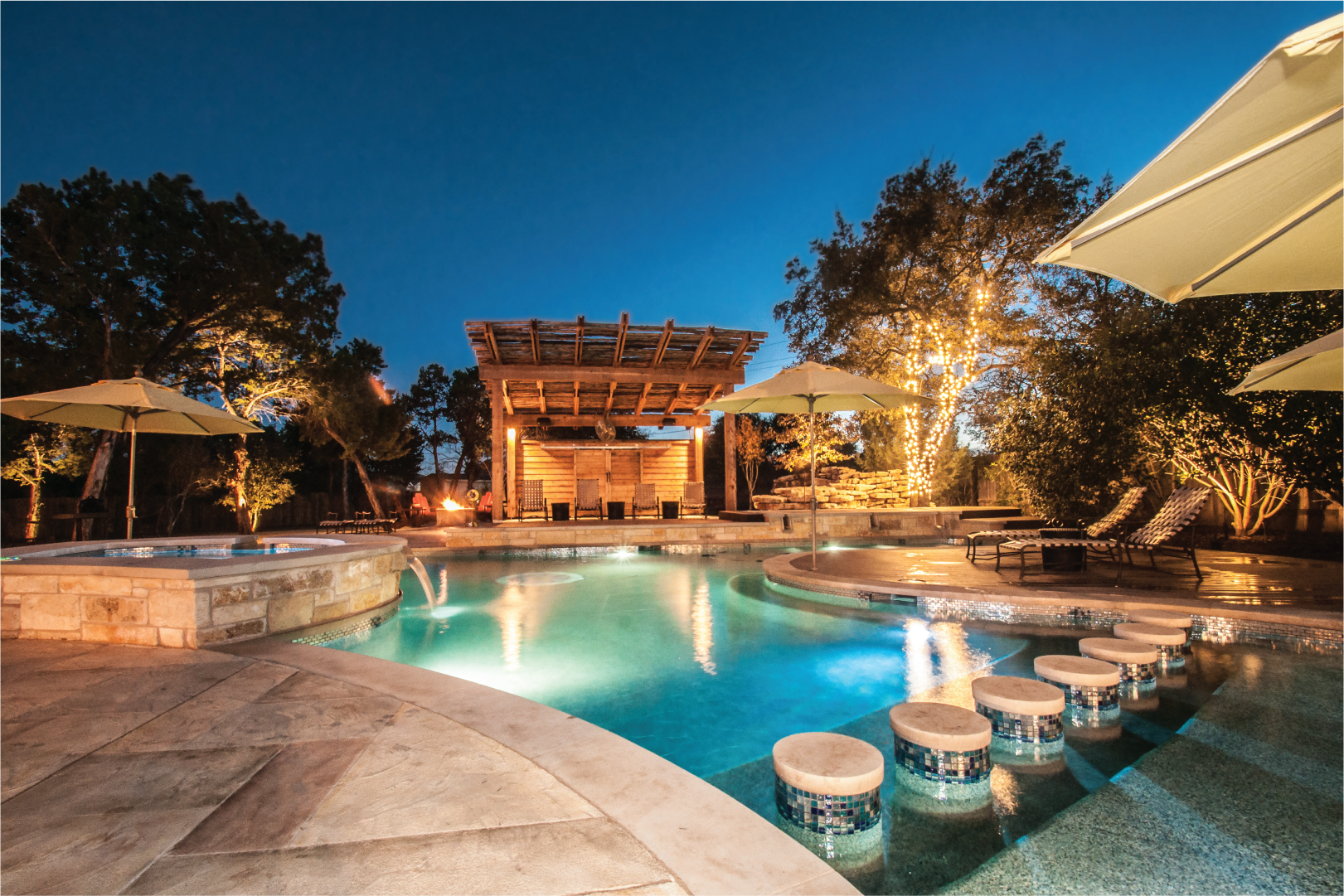 A beautiful back yard at night with lighting, a crystal clear pool, gazebo, jetted tub, and stone landscaping..