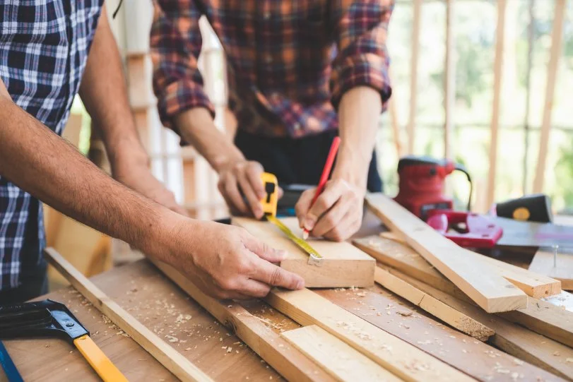 A close-in pphoto of two people measuring and marking wood while building a wood craft.