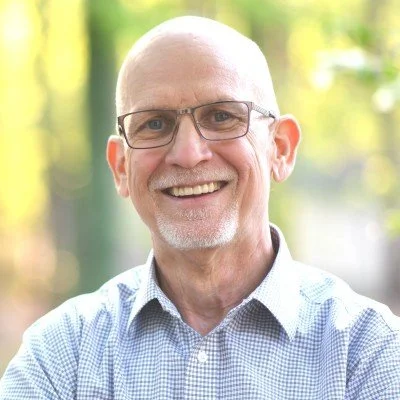 A smiling middle-aged man with glasses, wearing a checked shirt, standing outdoors with blurred green trees in the background.