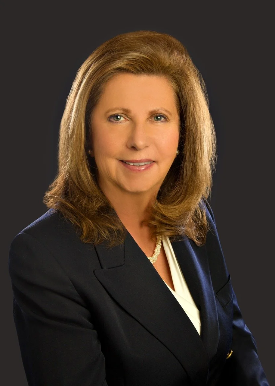 A professional headshot of a woman with brown hair, wearing a navy blazer, white blouse, pearl necklace, and pearl earrings, against a dark background.