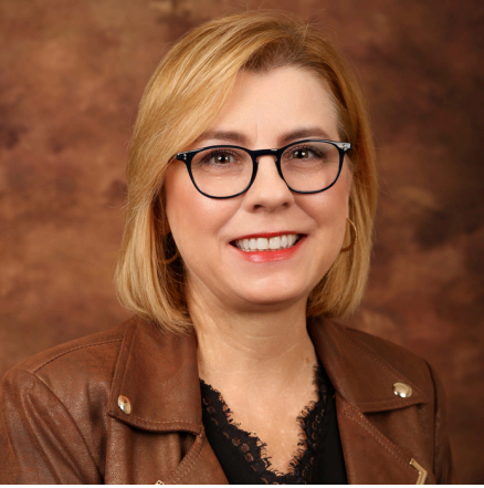 Portrait of a woman with blonde hair, glasses, and a brown leather jacket, smiling against a brown background.