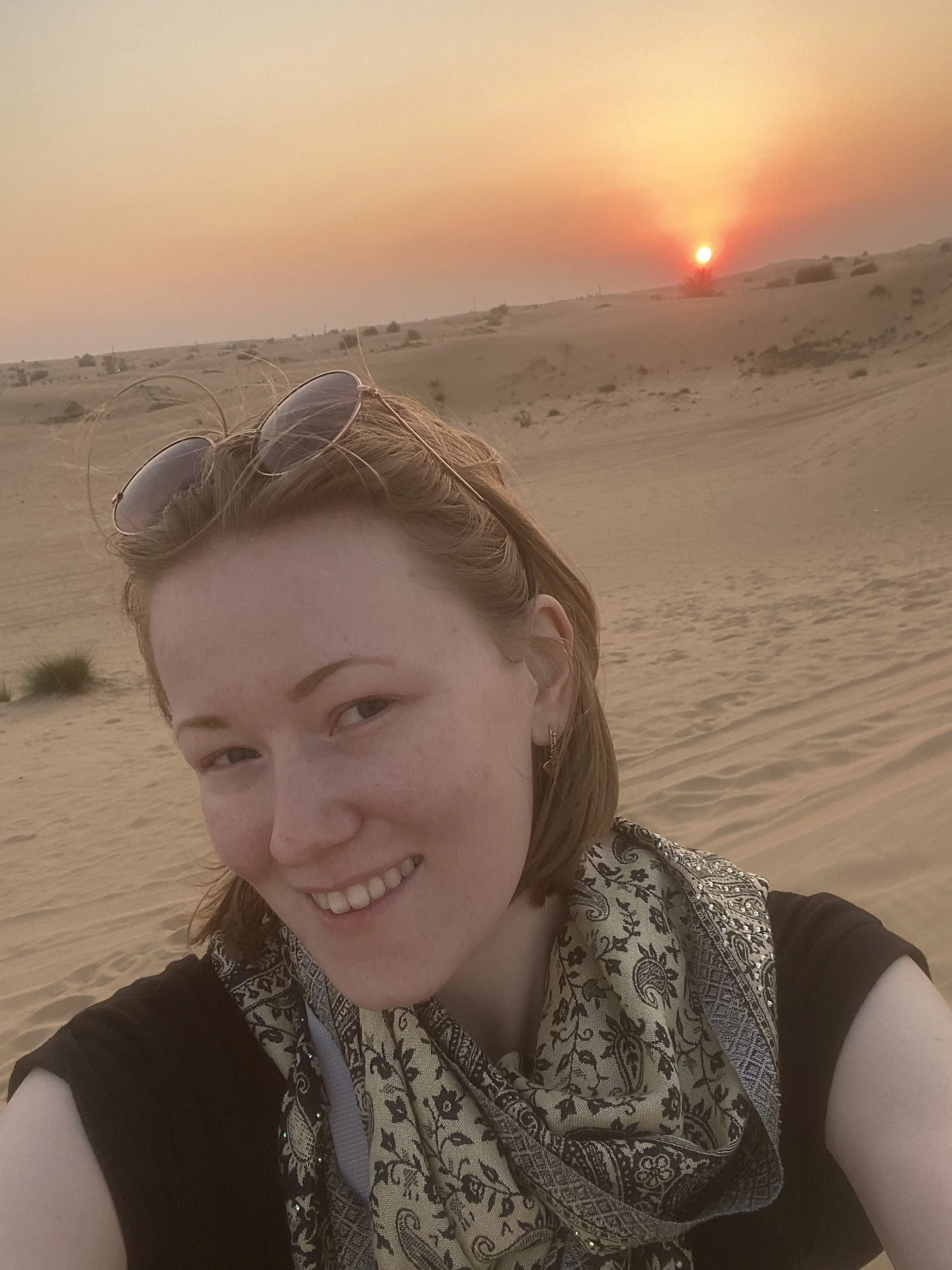 A woman taking a selfie in a desert at sunset, wearing sunglasses on her head and a paisley shirt.