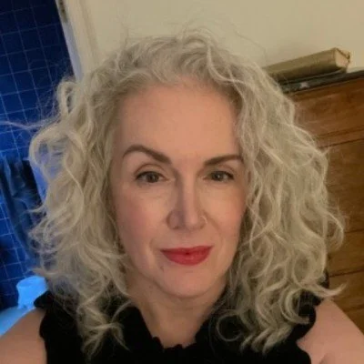 A woman with curly gray hair and red lipstick smiling indoors, wearing a black top, with a wooden dresser and window in the background.