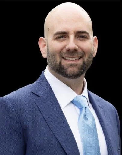 A bald man with a beard dressed in a blue suit, white shirt, and light blue tie, smiling against a black background.