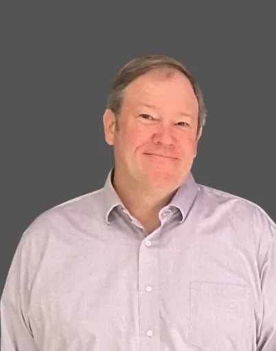 A middle-aged man with light skin and ginger hair, smiling gently, wearing a light purple button-up shirt, against a gray background.