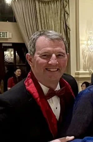 A smiling man in formal attire with a tuxedo, red bow tie, and red scarf, attending a social event in a decorated indoor setting.