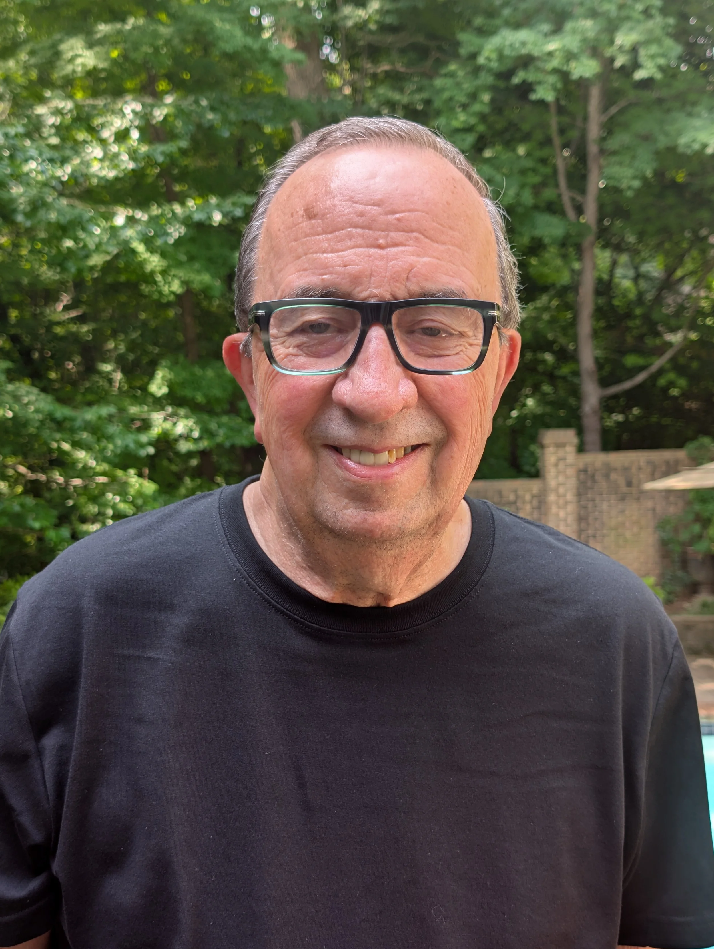A smiling man with glasses outdoors in front of green trees and a brick wall.