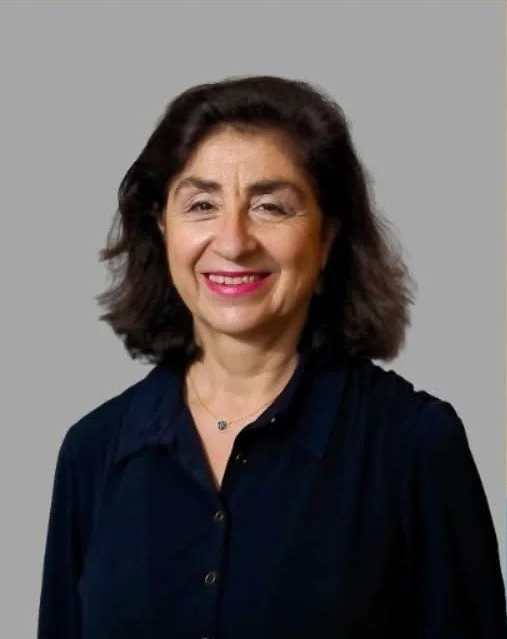 A woman with shoulder-length dark hair, smiling, wearing a dark blouse and a necklace, standing against a plain gray background.