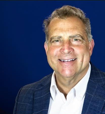A smiling middle-aged man with gray hair, wearing a dark blue blazer and white shirt, posing against a solid blue background.
