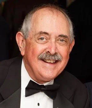 A smiling man with gray hair and mustache wearing a tuxedo with a bowtie.