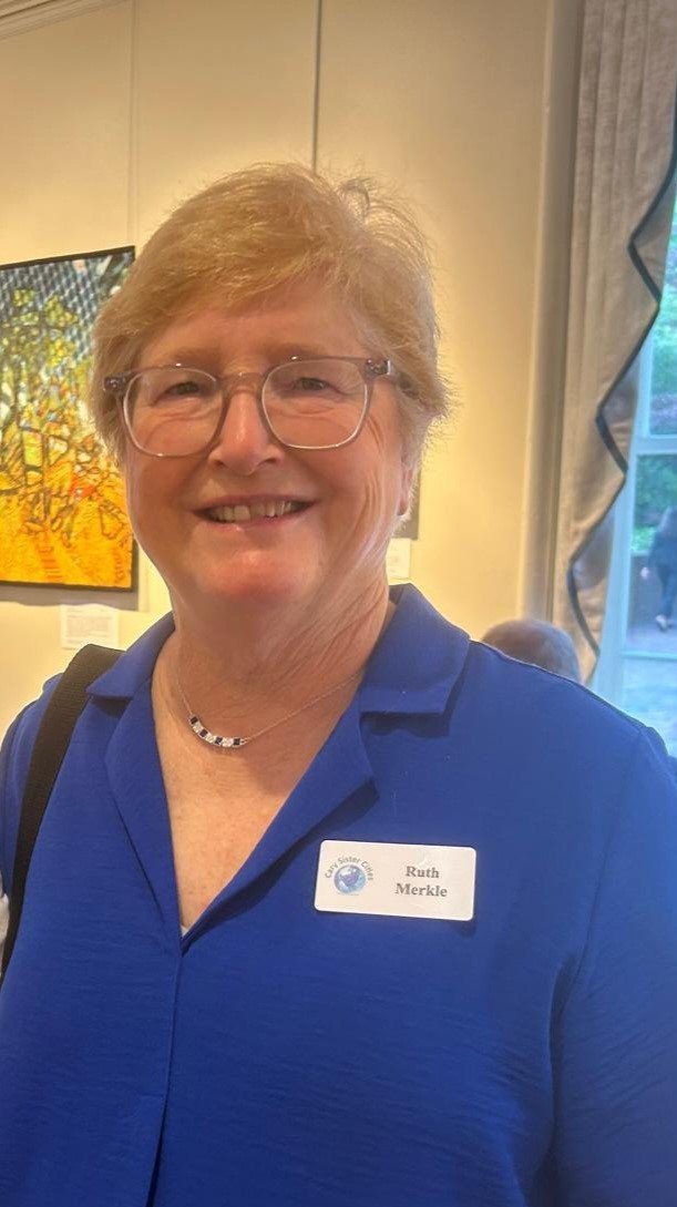 A smiling woman with short light brown hair and glasses, wearing a blue shirt and a name tag that reads 'Ruth Merkle', standing indoors near a window with curtains and artwork in the background.