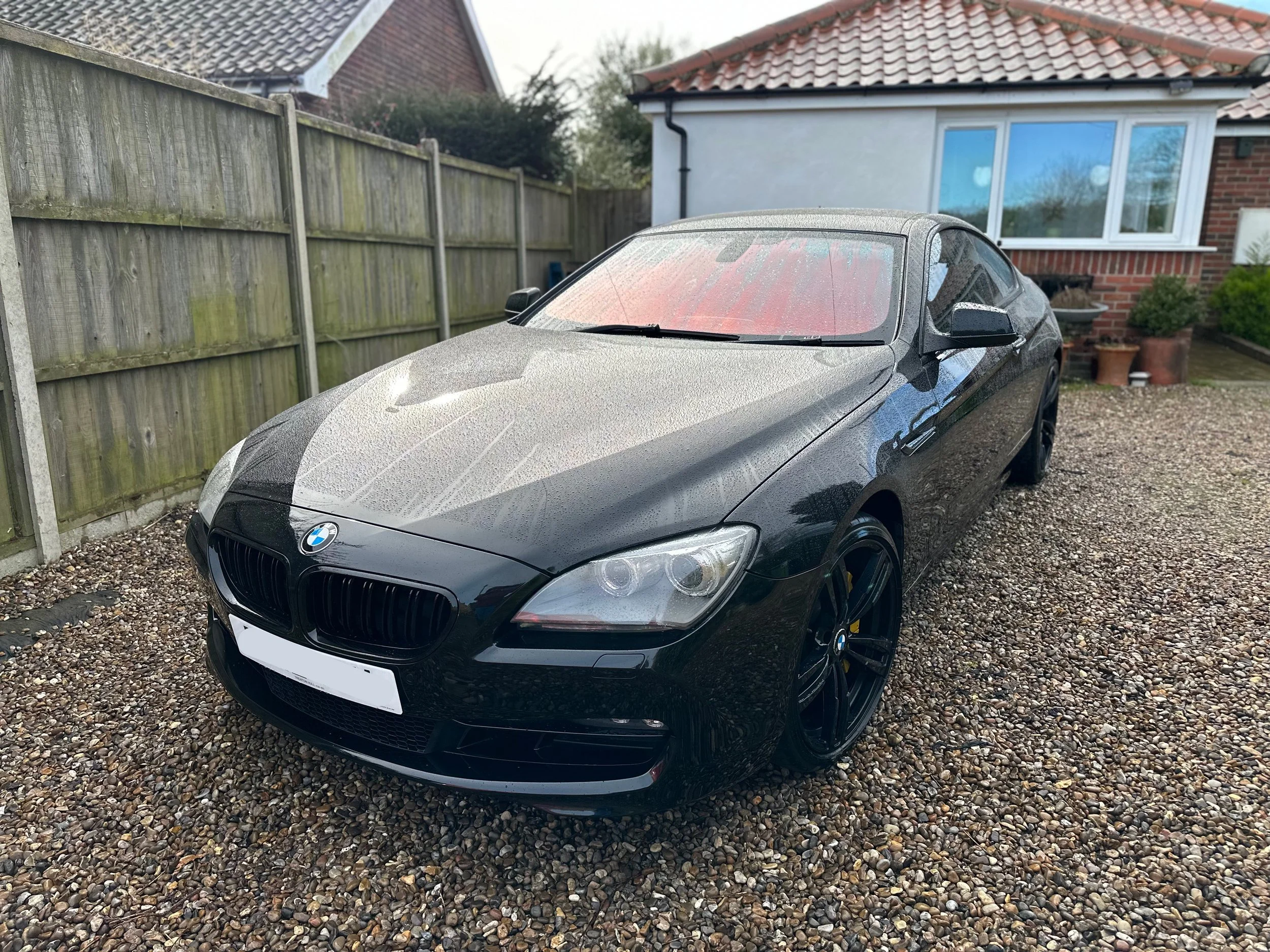 Black BMW sports car parked on a gravel driveway in front of a house with a white and brick exterior and a large window.