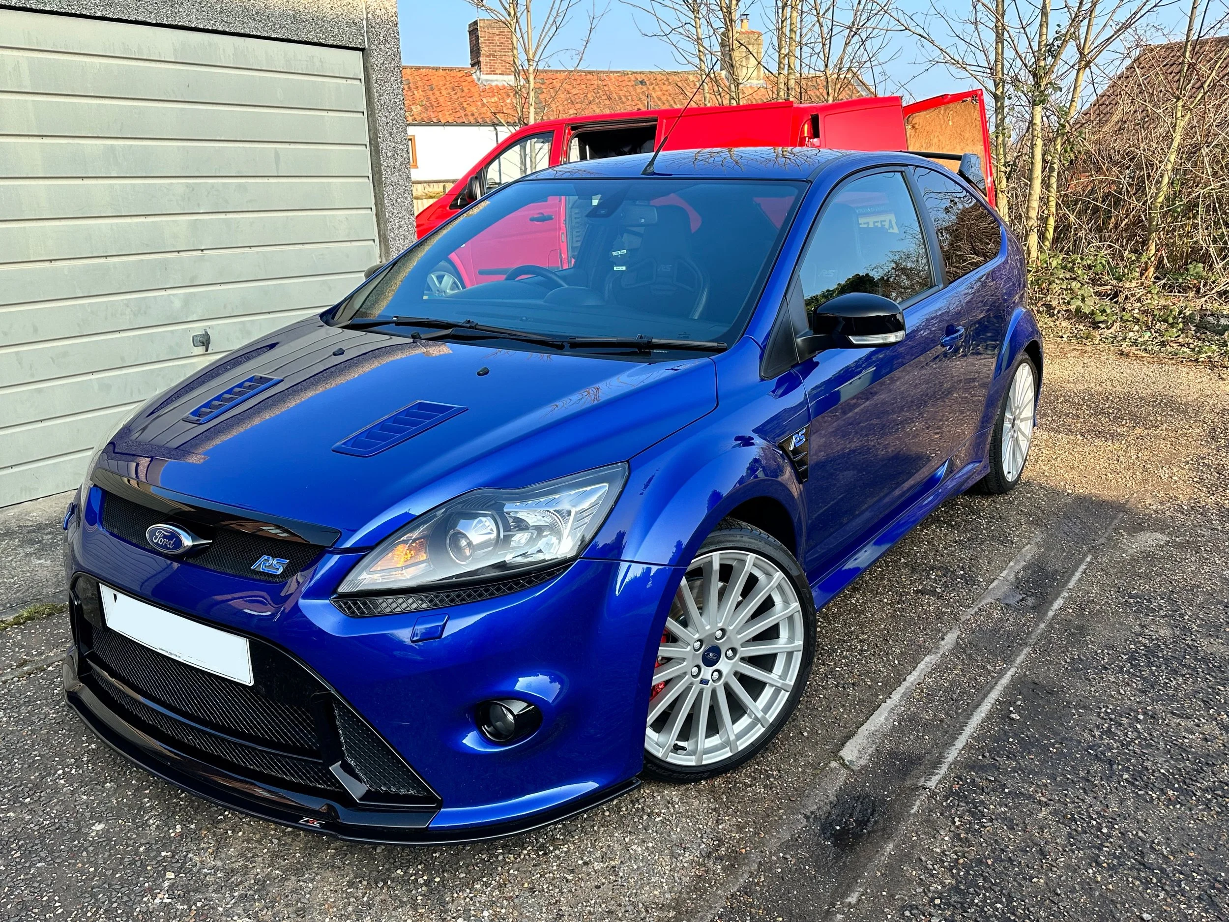 A blue Ford Focus RS hatchback parked outdoors on a gravel surface with a red van in the background and trees with no leaves.