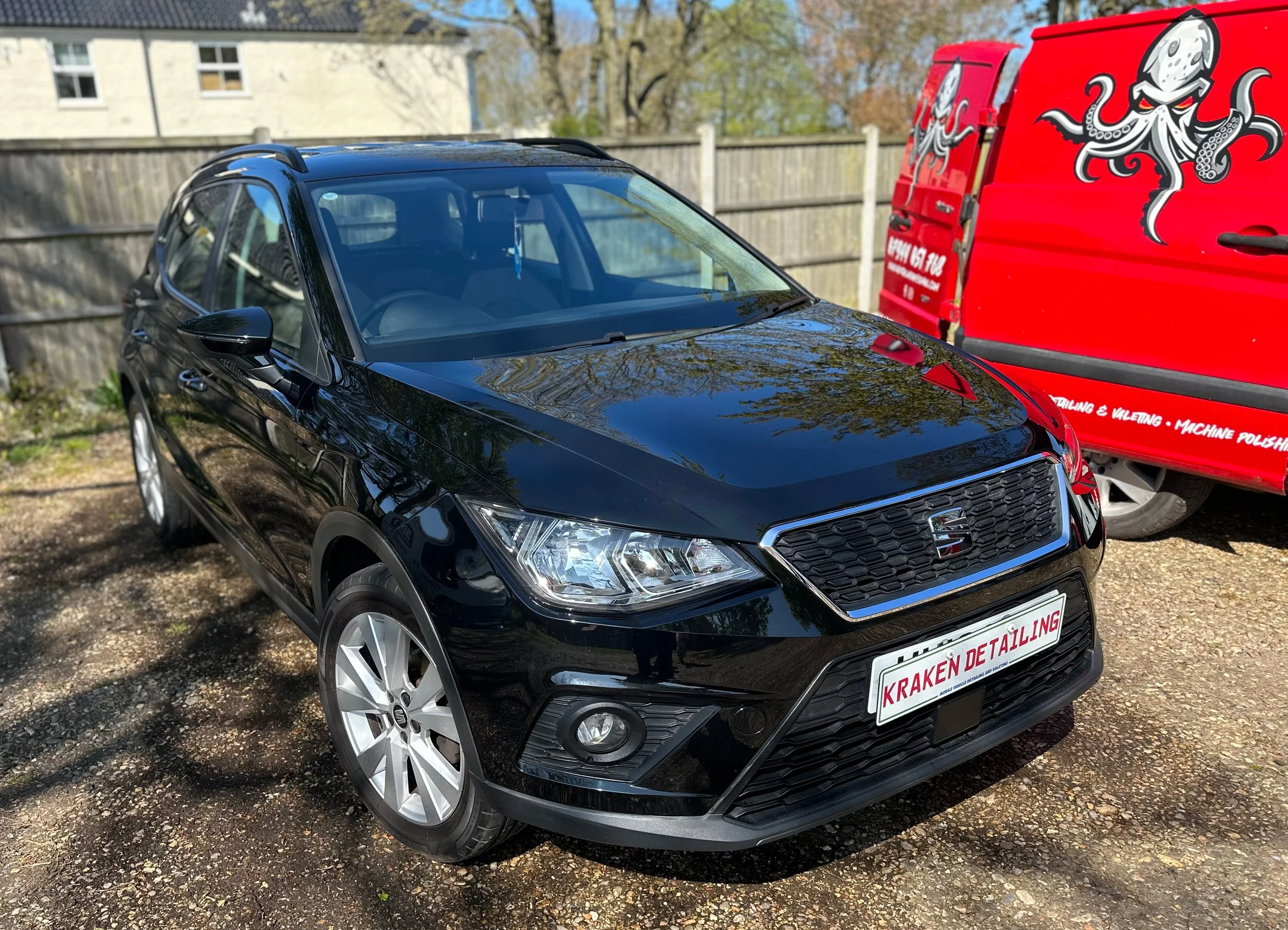 Black SEAT hatchback car parked on a dirt surface next to a red van with octopus artwork, fence, trees, and buildings in the background.