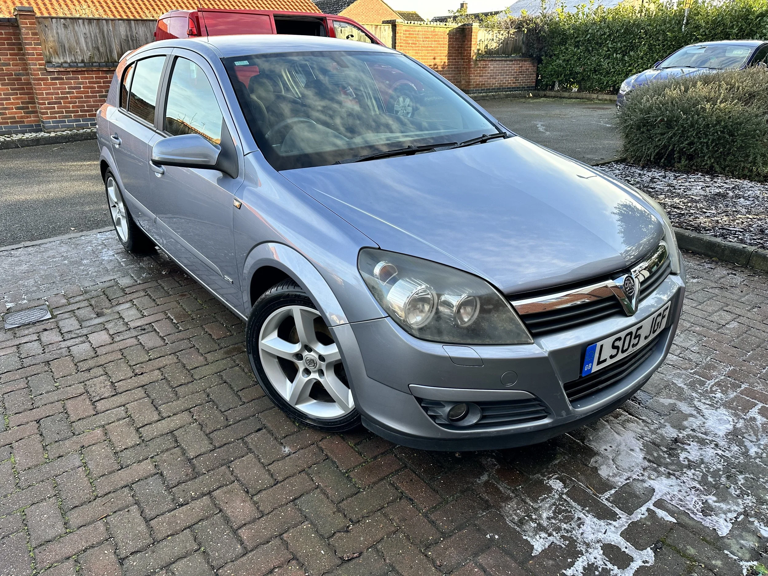 A silver Vauxhall Astra hatchback parked on a brick driveway with a brick wall and bushes in the background.