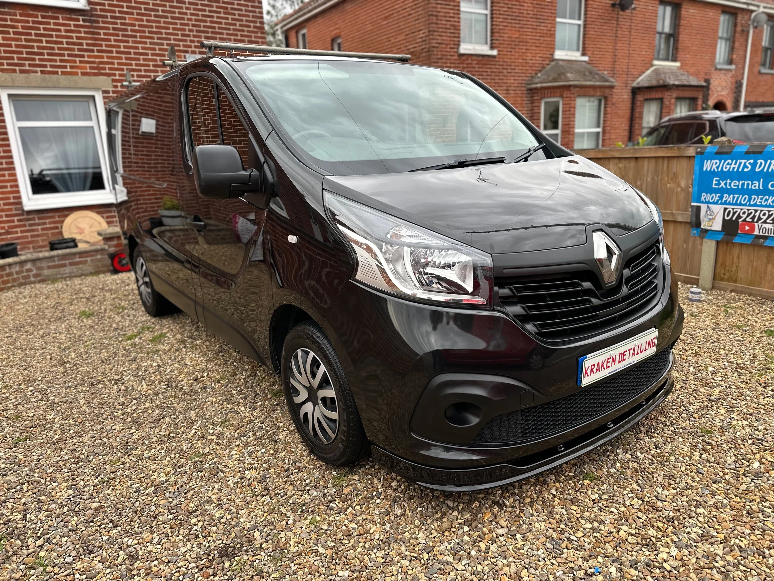 Black Renault van parked on gravel driveway in front of brick house with a window and a wooden fence.